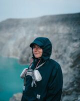 Kizzi standing at the top of Mount Ijen with the blue lake in the background wearing a gas mask around her neck